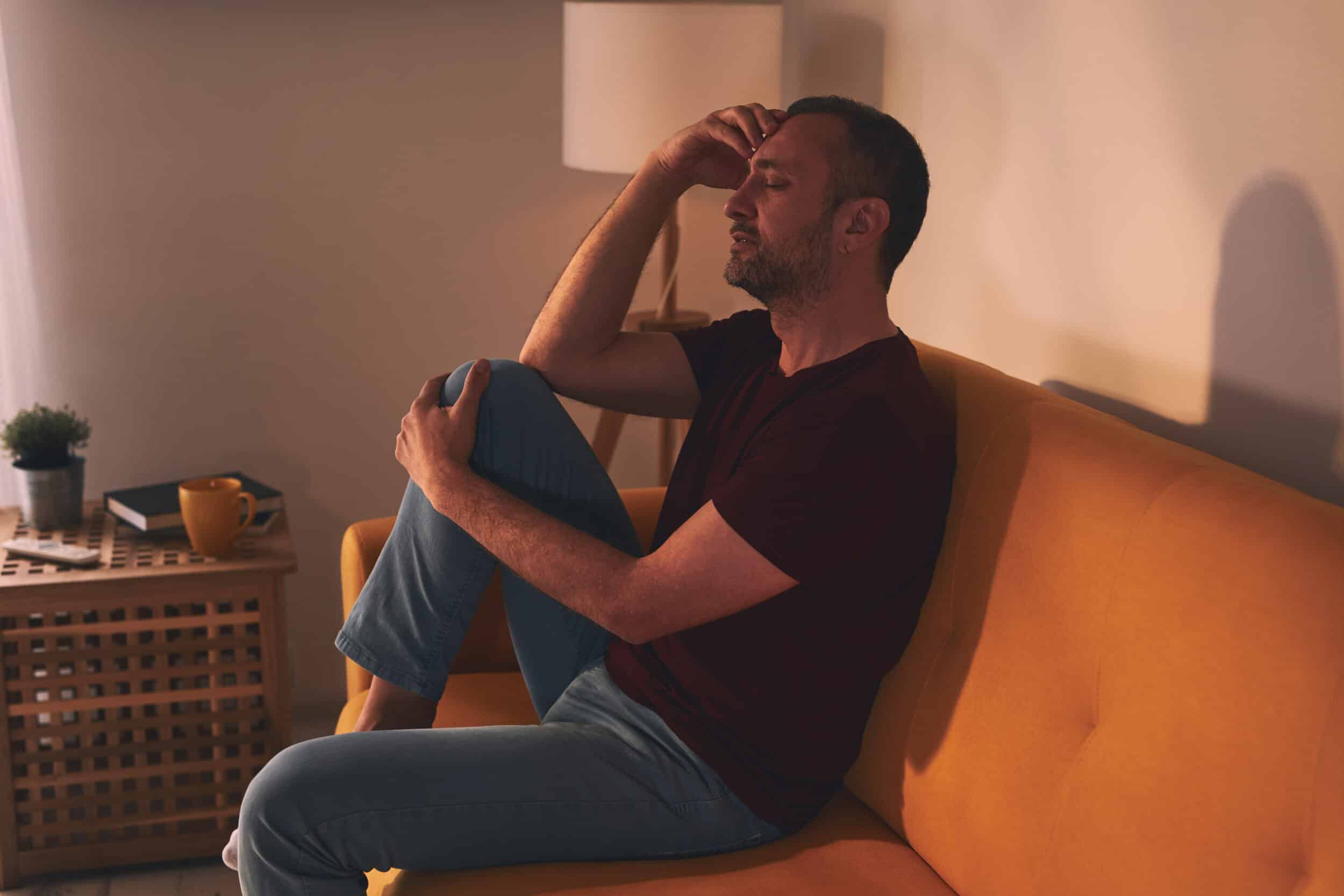 Man with headache and knee pain sitting on a sofa at home.